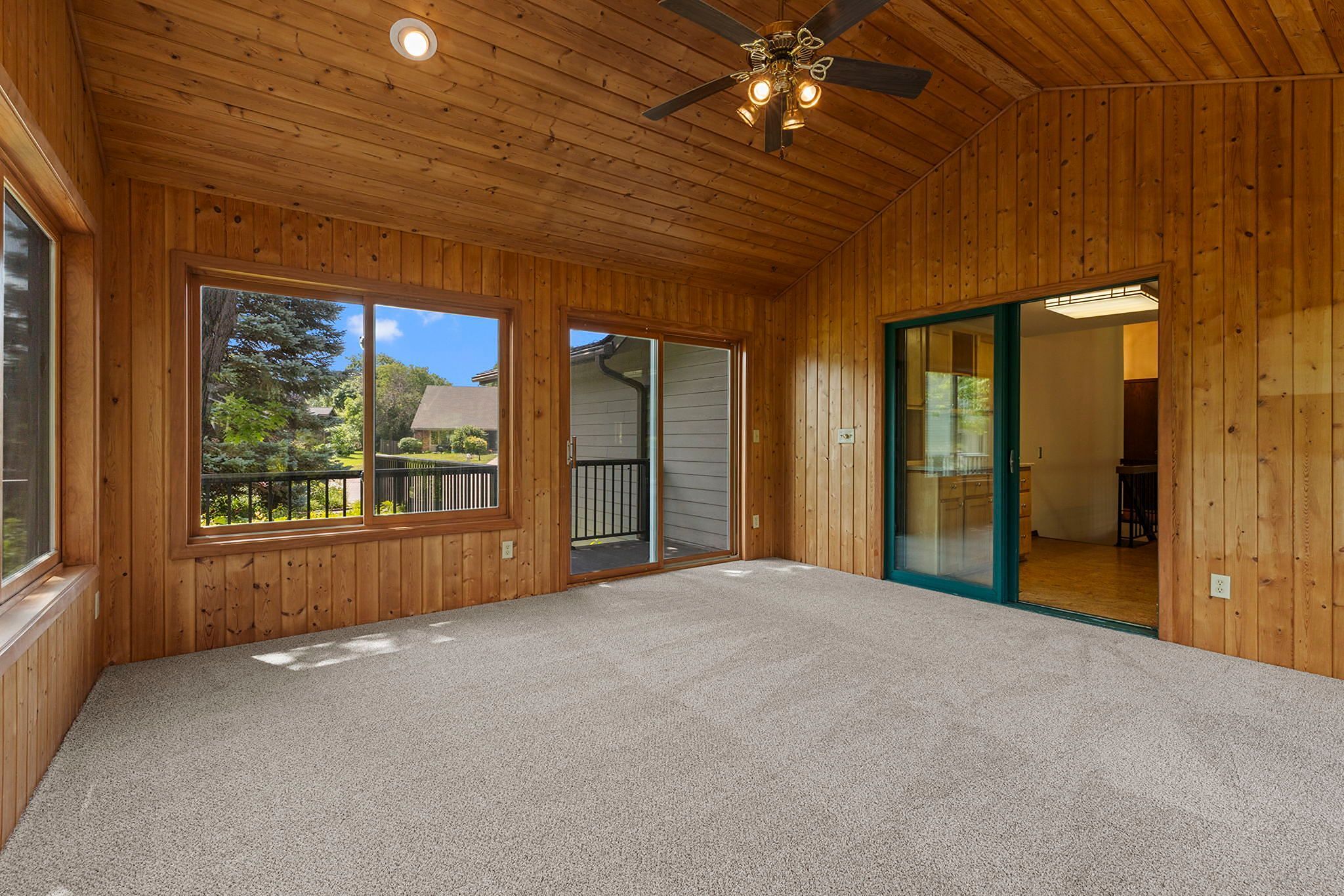 A sunroom with wood-paneled walls and ceiling, large windows, a ceiling fan with lights, carpeted floor, and a sliding glass door overlooking a patio and greenery outside.