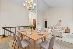A modern dining area with a light wood table set for six, gray cushioned chairs, and a floral centerpiece, opens to a bright living room with white sofas, a coffee table, and decorative accents under a high ceiling.