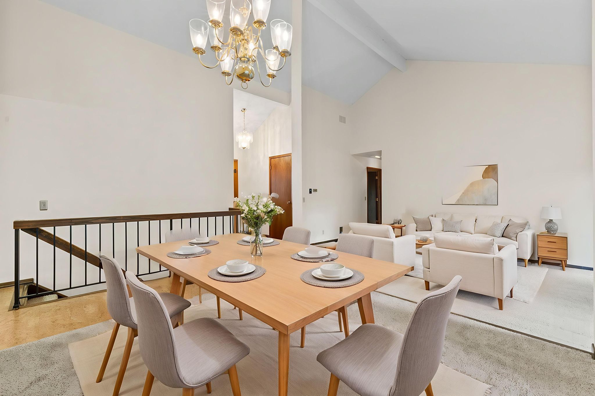 A modern dining area with a light wood table set for six, gray cushioned chairs, and a floral centerpiece, opens to a bright living room with white sofas, a coffee table, and decorative accents under a high ceiling.