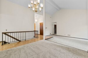 Spacious living area with light carpet, vaulted ceiling, brass chandelier, white walls, and a black metal railing overlooking a lower stairway. Wood door and hallway are visible in the background.