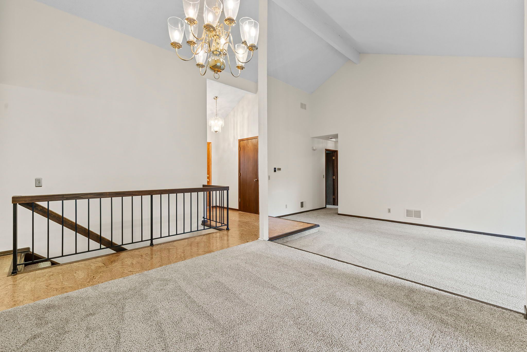 Spacious living area with light carpet, vaulted ceiling, brass chandelier, white walls, and a black metal railing overlooking a lower stairway. Wood door and hallway are visible in the background.