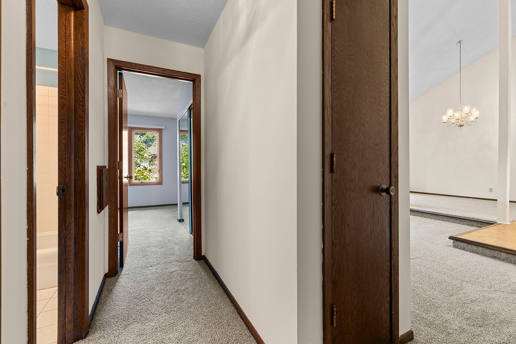 A hallway with beige carpet, wooden doors, and white walls leads to a bedroom and bathroom; a sunlit living area with a chandelier is partially visible to the right.