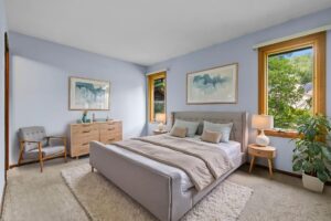 A modern bedroom with a gray bed, beige bedding, wooden dresser, chair, two large windows, a potted plant, and light blue walls decorated with abstract art.