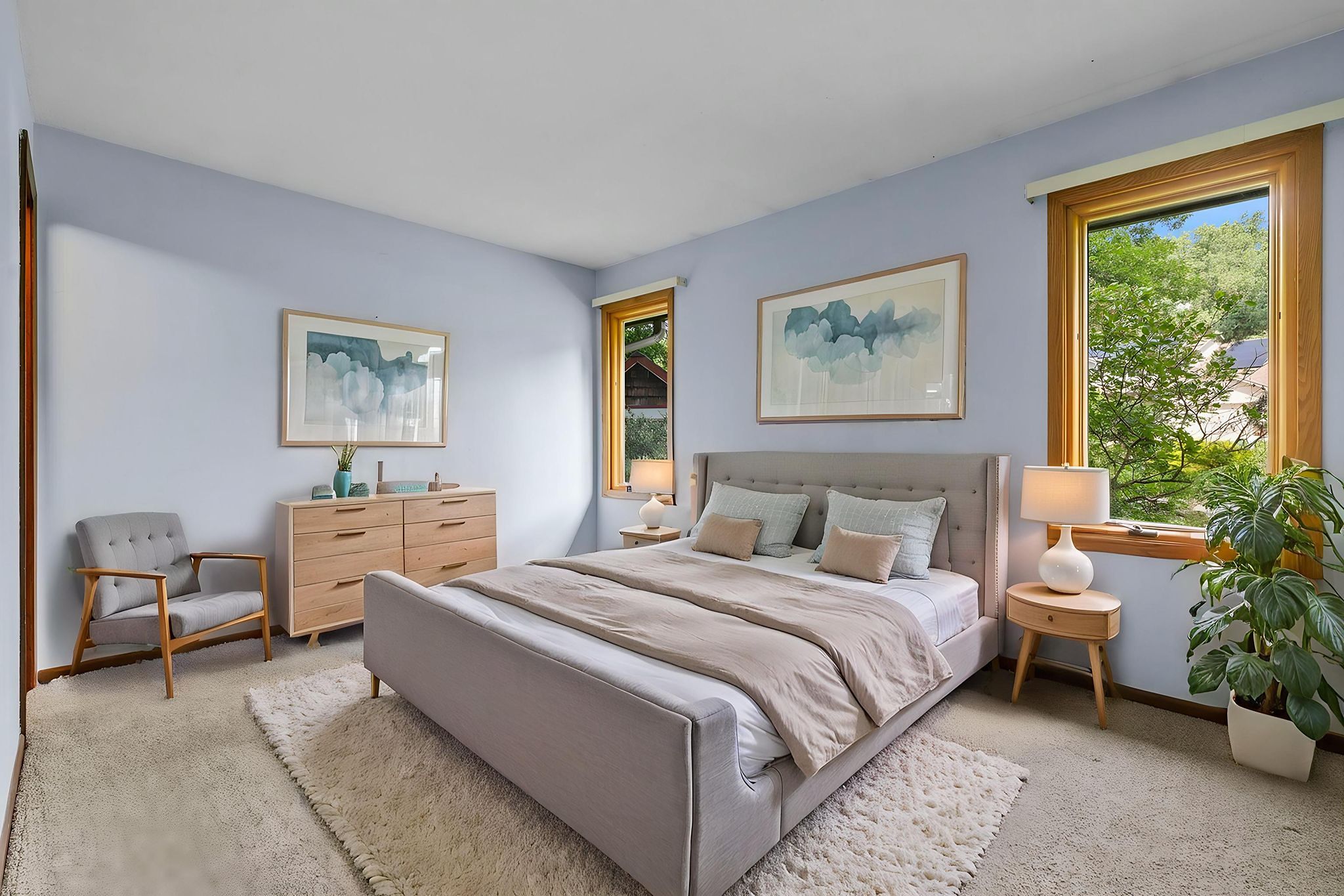 A modern bedroom with a gray bed, beige bedding, wooden dresser, chair, two large windows, a potted plant, and light blue walls decorated with abstract art.