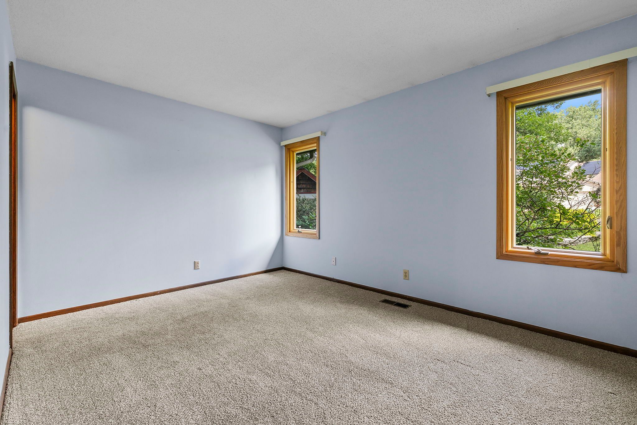 Empty room with light blue walls, beige carpet, two windows with wood trim, and a view of greenery and houses outside. The room has a vent on the floor and electrical outlets on the walls.
