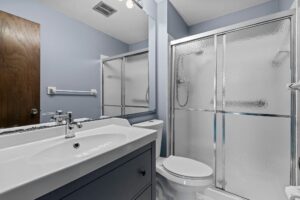 A modern bathroom with a white sink, a blue vanity, a toilet, and a glass shower enclosure. The walls are light blue and there is a wooden door and a large mirror above the sink.