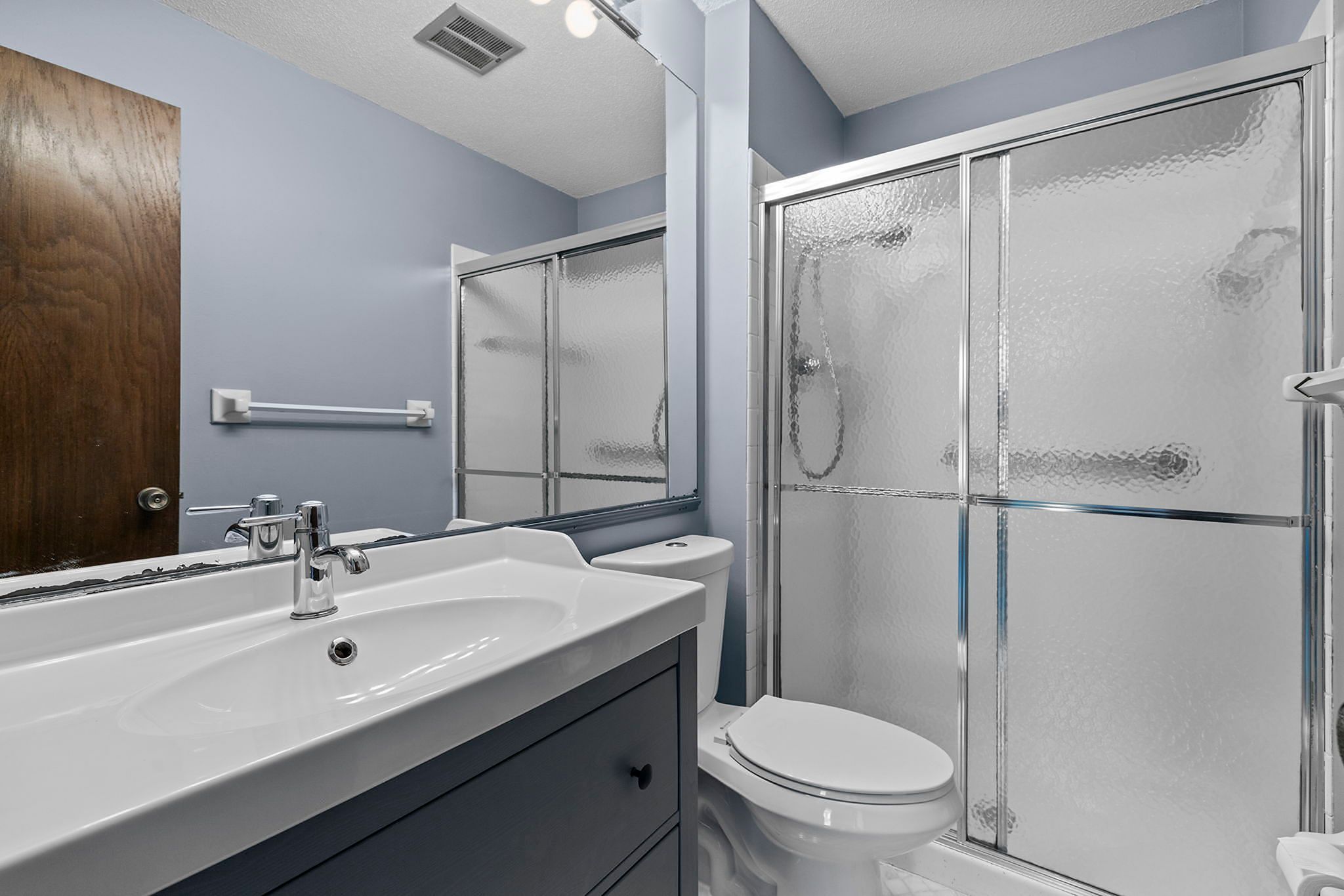 A modern bathroom with a white sink, a blue vanity, a toilet, and a glass shower enclosure. The walls are light blue and there is a wooden door and a large mirror above the sink.