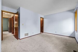 Empty bedroom with beige carpet, light blue walls, wooden trim, two open wooden doors leading to a hallway, and a window letting in natural light.