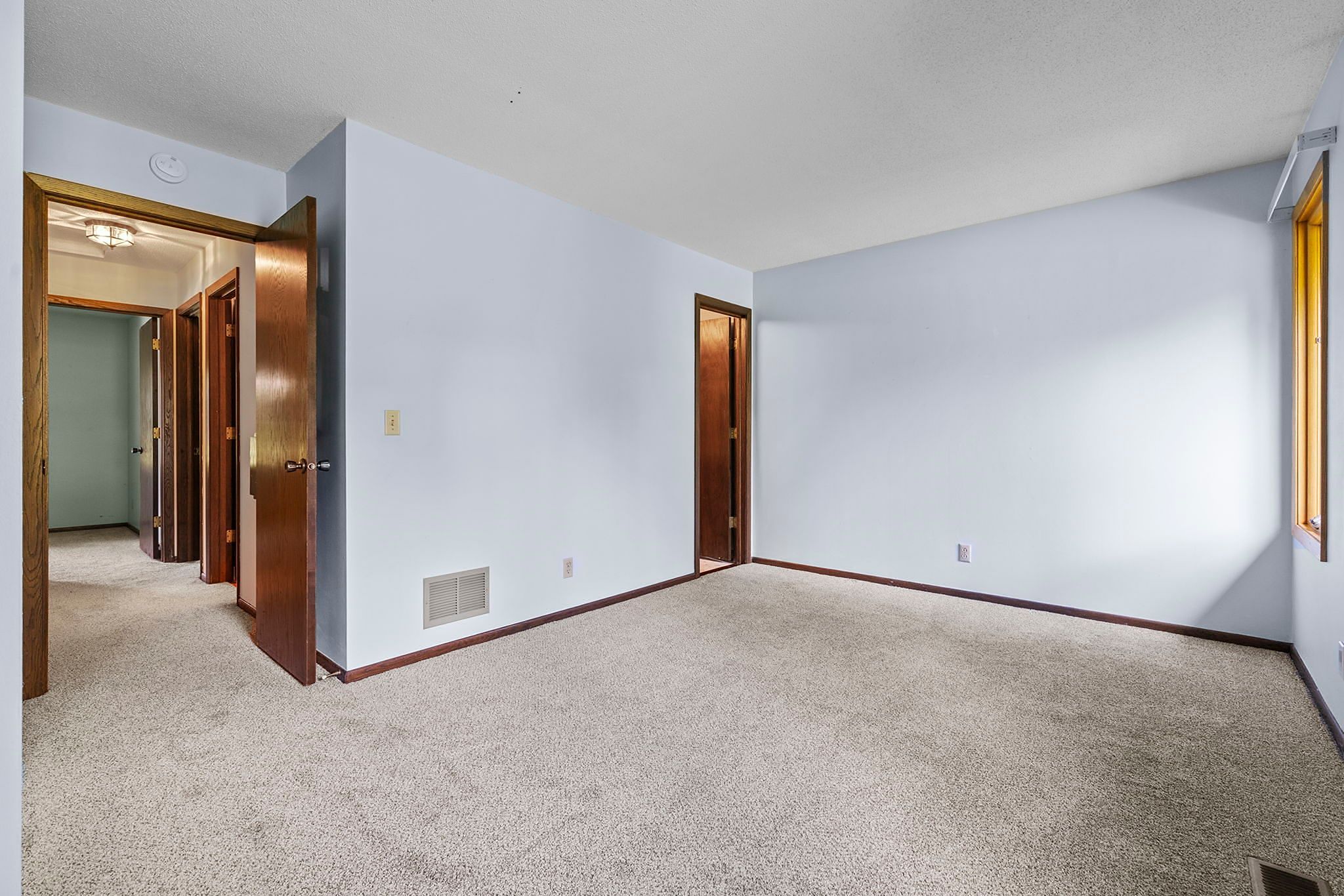 Empty bedroom with beige carpet, light blue walls, wooden trim, two open wooden doors leading to a hallway, and a window letting in natural light.