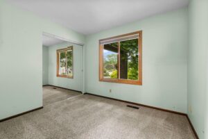 Empty room with light green walls, beige carpet, large window with wood trim showing trees outside, and a mirrored closet door reflecting the room and window.