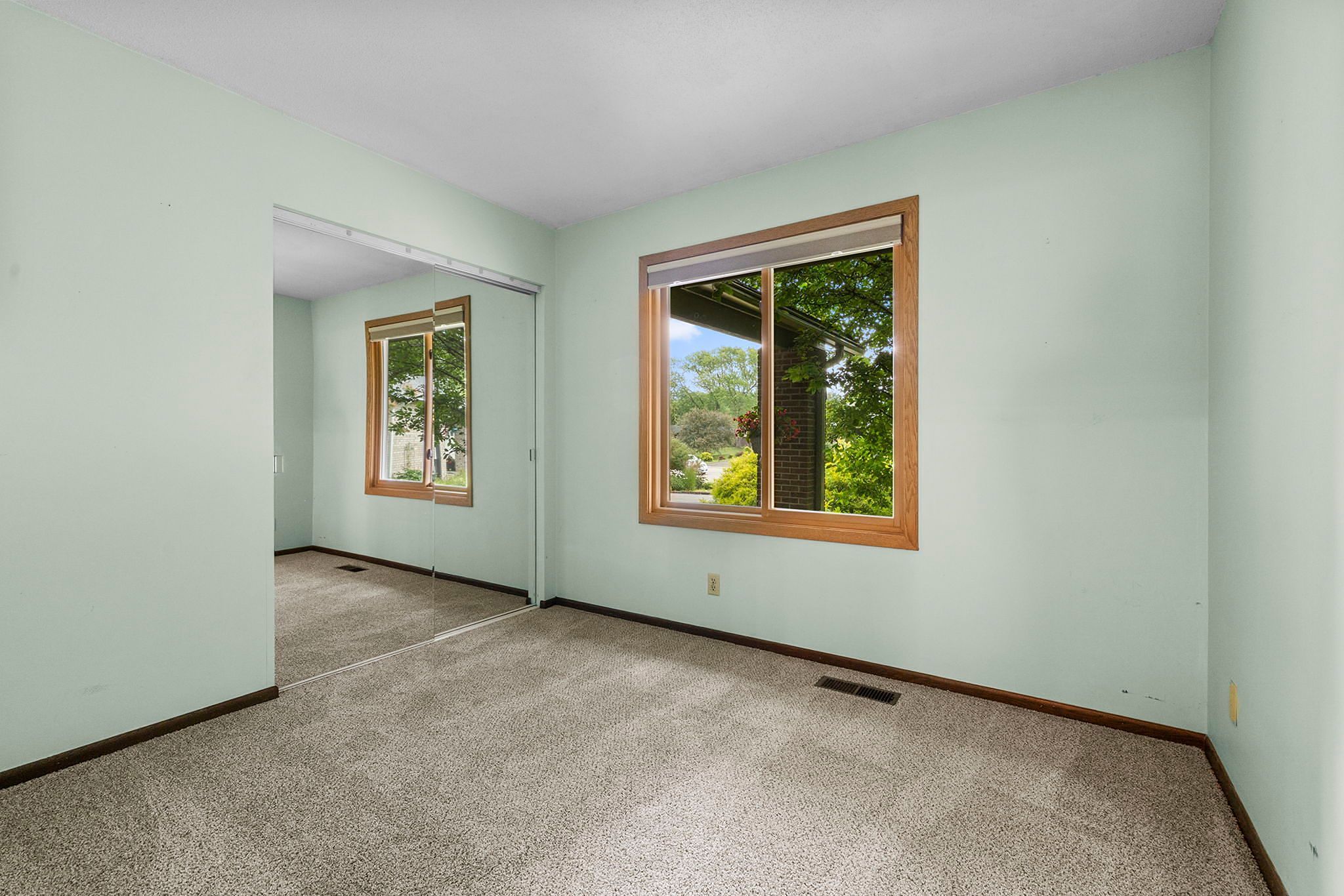 Empty room with light green walls, beige carpet, large window with wood trim showing trees outside, and a mirrored closet door reflecting the room and window.