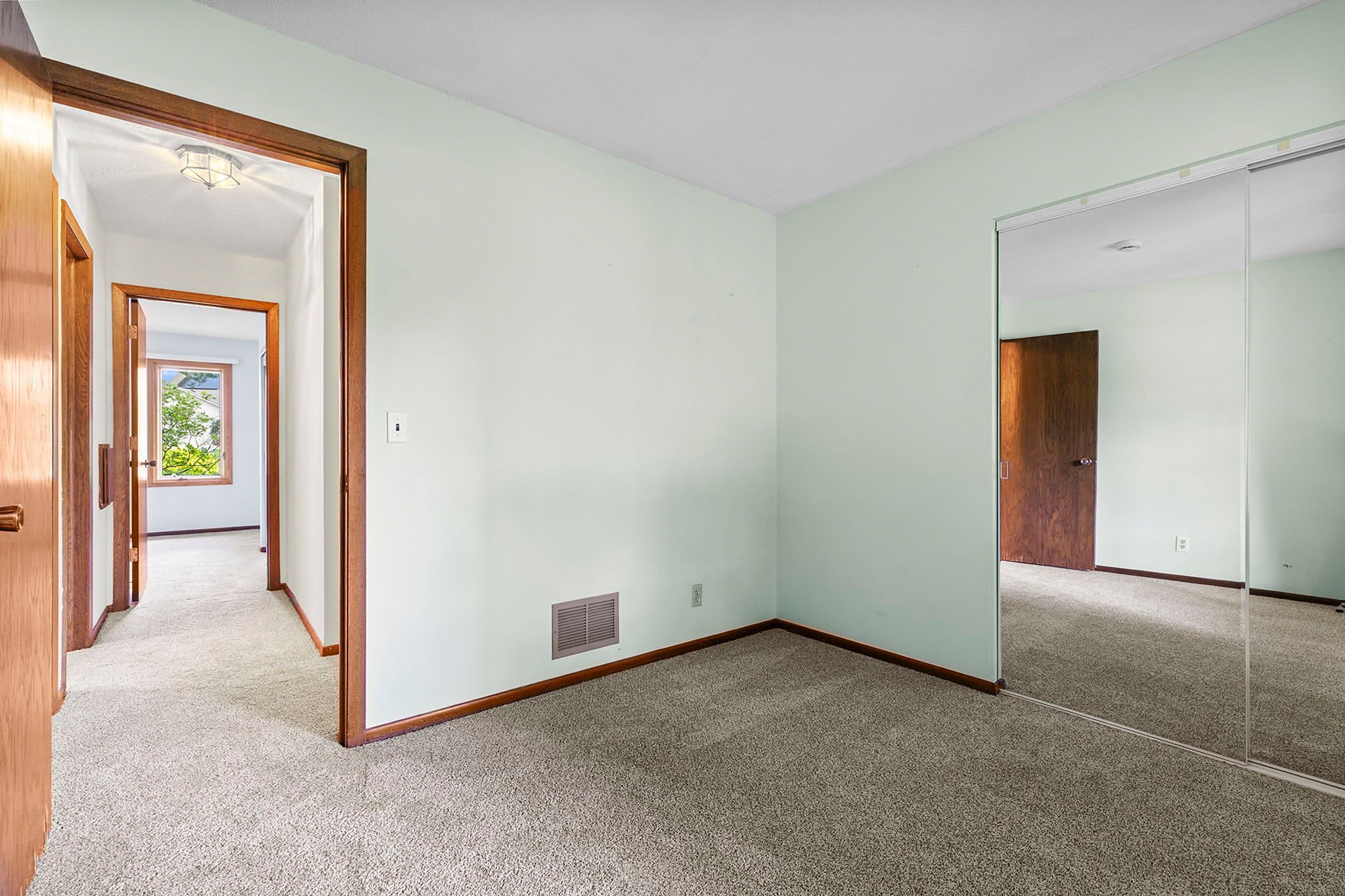 Empty carpeted bedroom with light green walls, wooden trim, mirrored closet doors, and open doorways leading to a hallway with additional rooms visible. Natural light comes from a window in the hall.