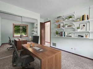 Modern home office with two wooden desks, office chairs, computers, and wall-mounted shelves holding books and plants. A large window offers a view of greenery outside, and mirrored closet doors reflect the room.