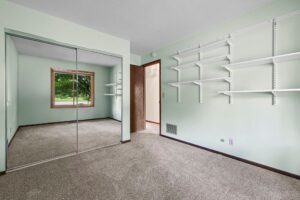 Empty room with light green walls, beige carpet, wall shelves, a large window, and mirrored closet doors reflecting the opposite wall and window view of green trees outside.
