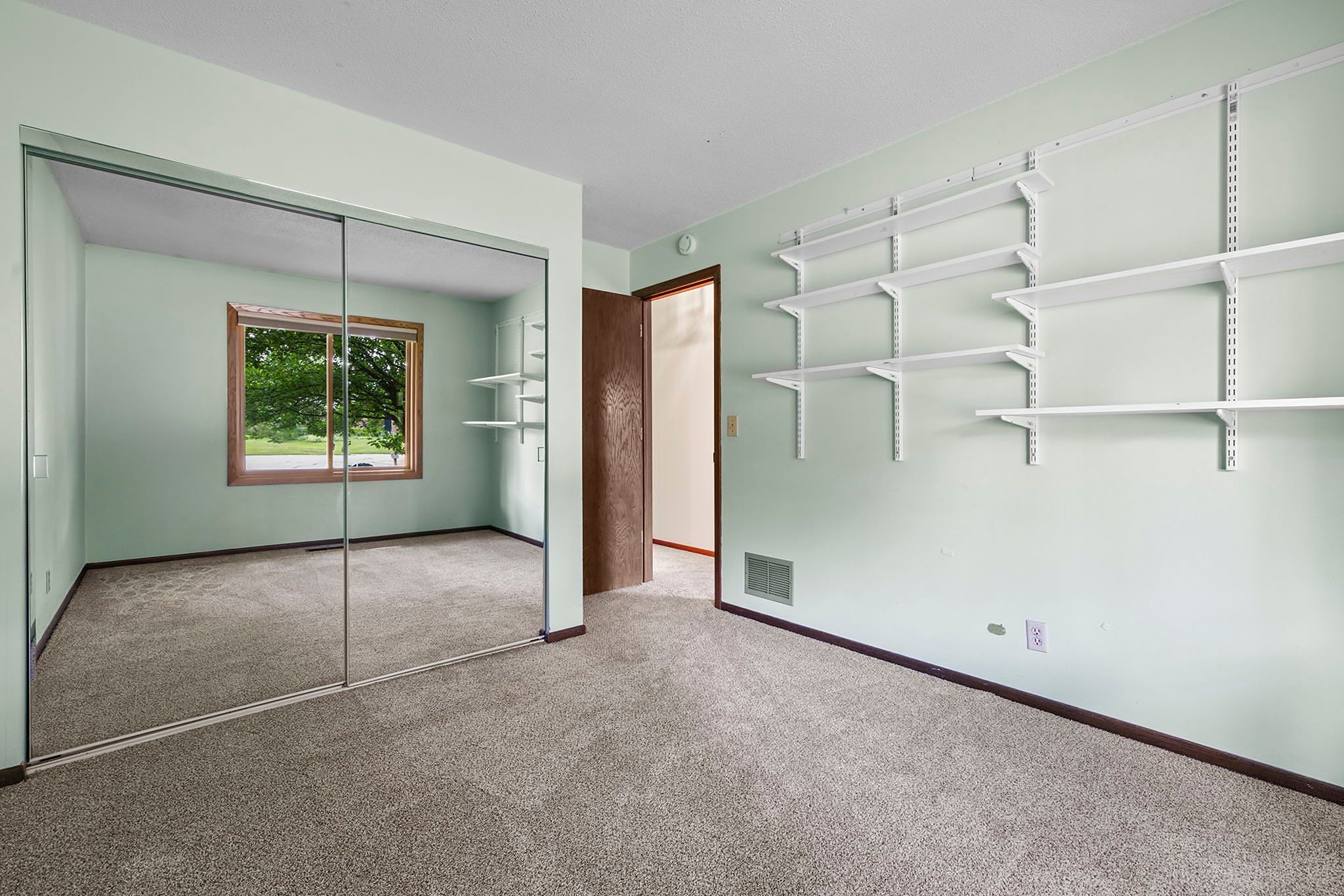 Empty room with light green walls, beige carpet, wall shelves, a large window, and mirrored closet doors reflecting the opposite wall and window view of green trees outside.