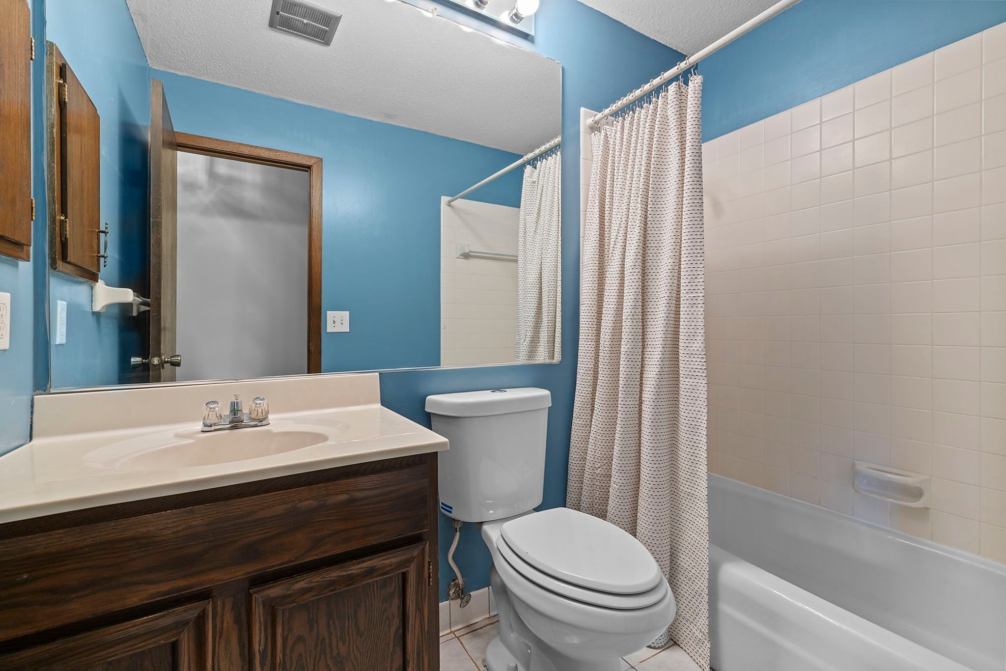 A bathroom with blue walls, a wooden vanity, white sink, mirror cabinet, toilet, bathtub with a white shower curtain, and tiled floor.