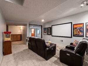 Home theater room with two brown leather recliners, a large projection screen, classic movie posters on the walls, and a popcorn machine in the corner. Carpeted floor and neutral walls create a cozy atmosphere.