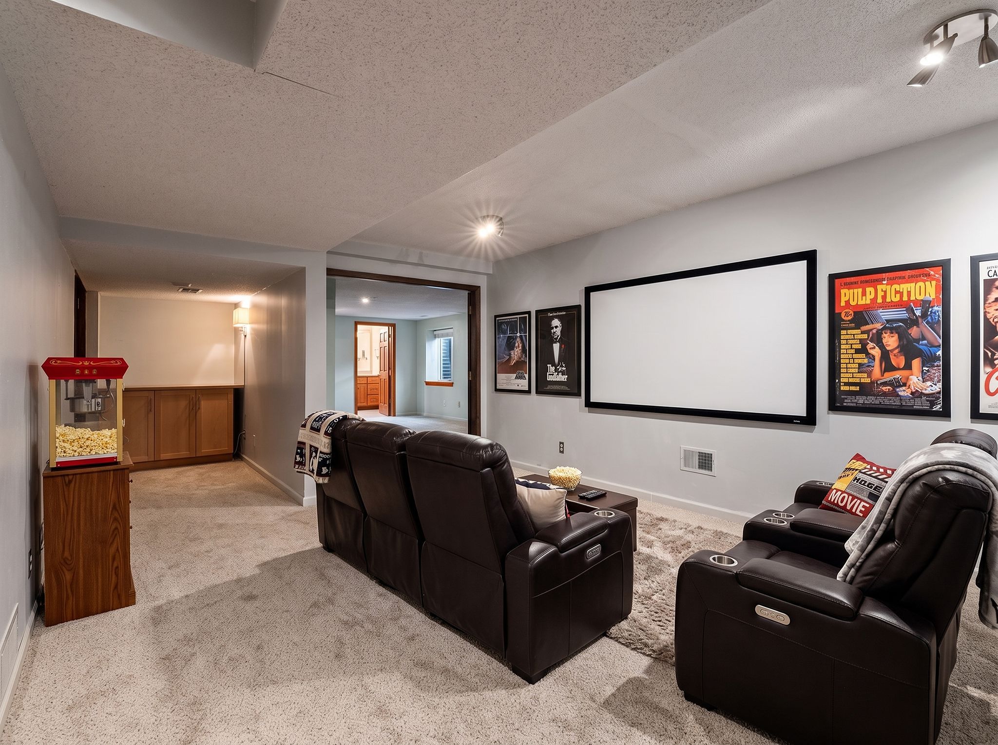 Home theater room with two brown leather recliners, a large projection screen, classic movie posters on the walls, and a popcorn machine in the corner. Carpeted floor and neutral walls create a cozy atmosphere.