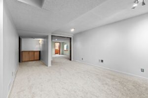 Spacious, carpeted basement room with white walls, recessed lighting, and built-in wooden cabinets in the corner. An open doorway leads to another room with wood accents.