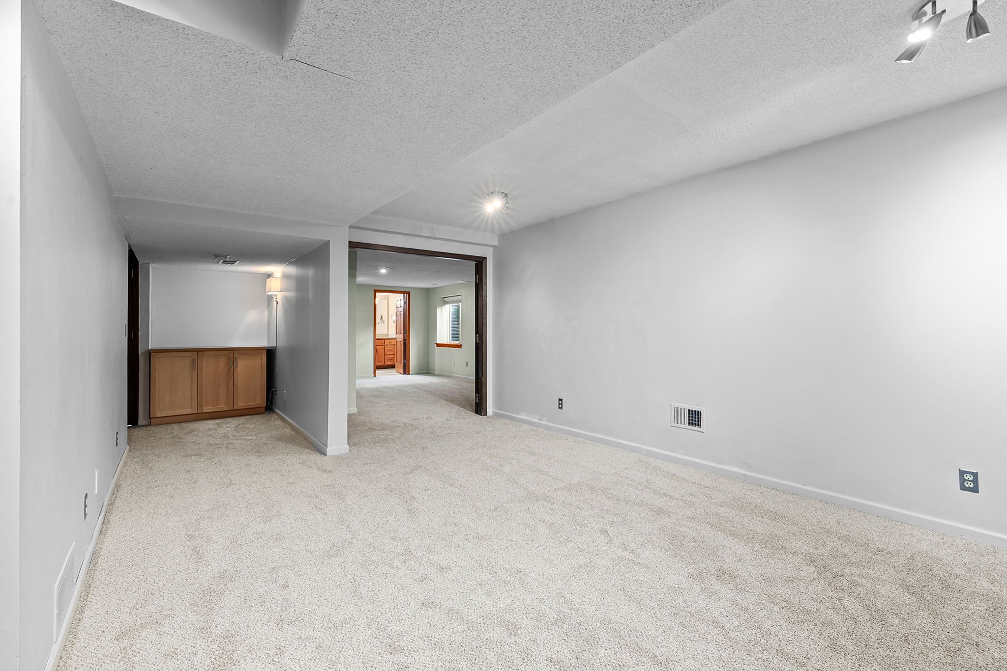 Spacious, carpeted basement room with white walls, recessed lighting, and built-in wooden cabinets in the corner. An open doorway leads to another room with wood accents.