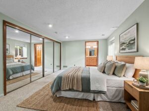 A cozy bedroom with a neatly made bed, wooden nightstand, lamp, large mirrored closet doors, and an open doorway leading to a bathroom. Soft lighting and neutral decor create a calm atmosphere.