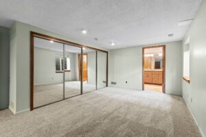 Spacious carpeted room with mirrored closet doors, light green walls, recessed lighting, and an attached bathroom with wooden cabinetry and a large mirror.