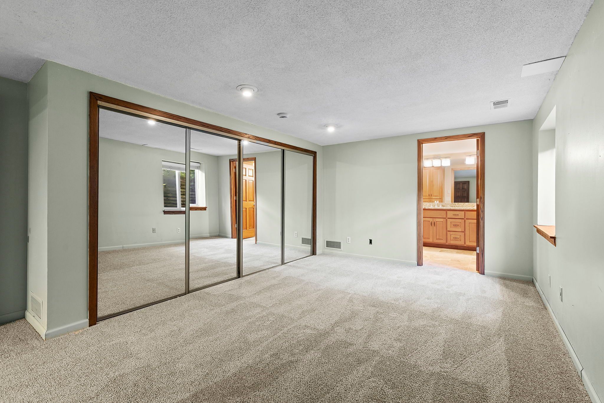 Spacious carpeted room with mirrored closet doors, light green walls, recessed lighting, and an attached bathroom with wooden cabinetry and a large mirror.
