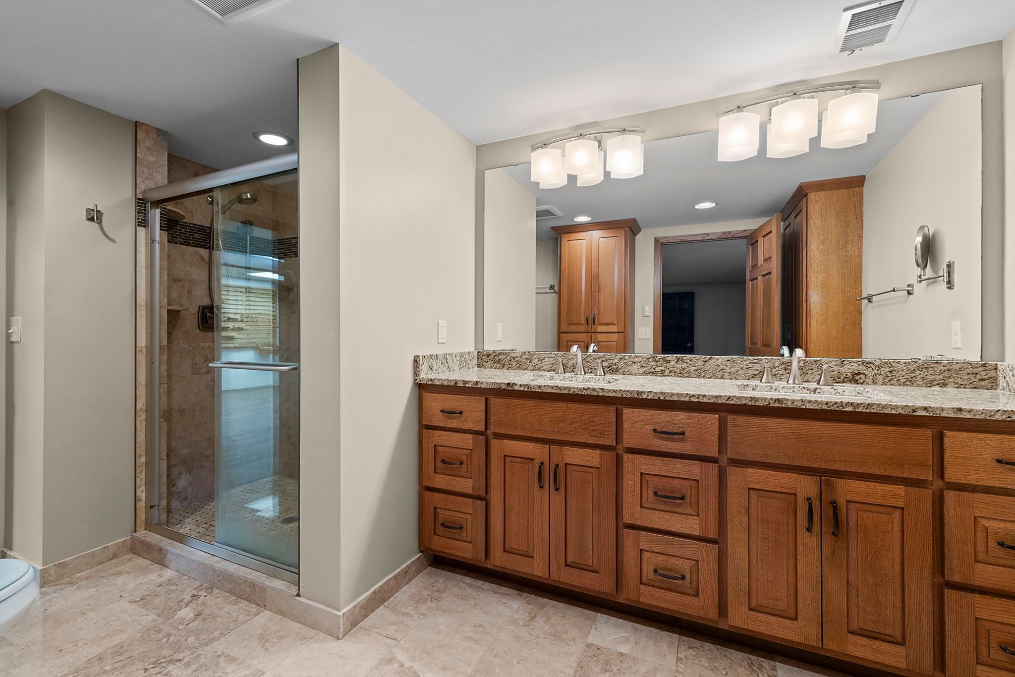 Modern bathroom with a double-sink wooden vanity, granite countertop, large mirror with lights, and a glass-enclosed walk-in shower. Marble floor tiles and neutral wall colors.