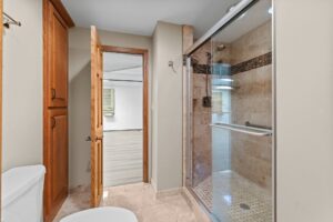 Modern bathroom with a glass-enclosed shower, tiled walls and floor, wooden cabinet, and toilet. An open wooden door leads to an adjacent room with light-colored flooring.