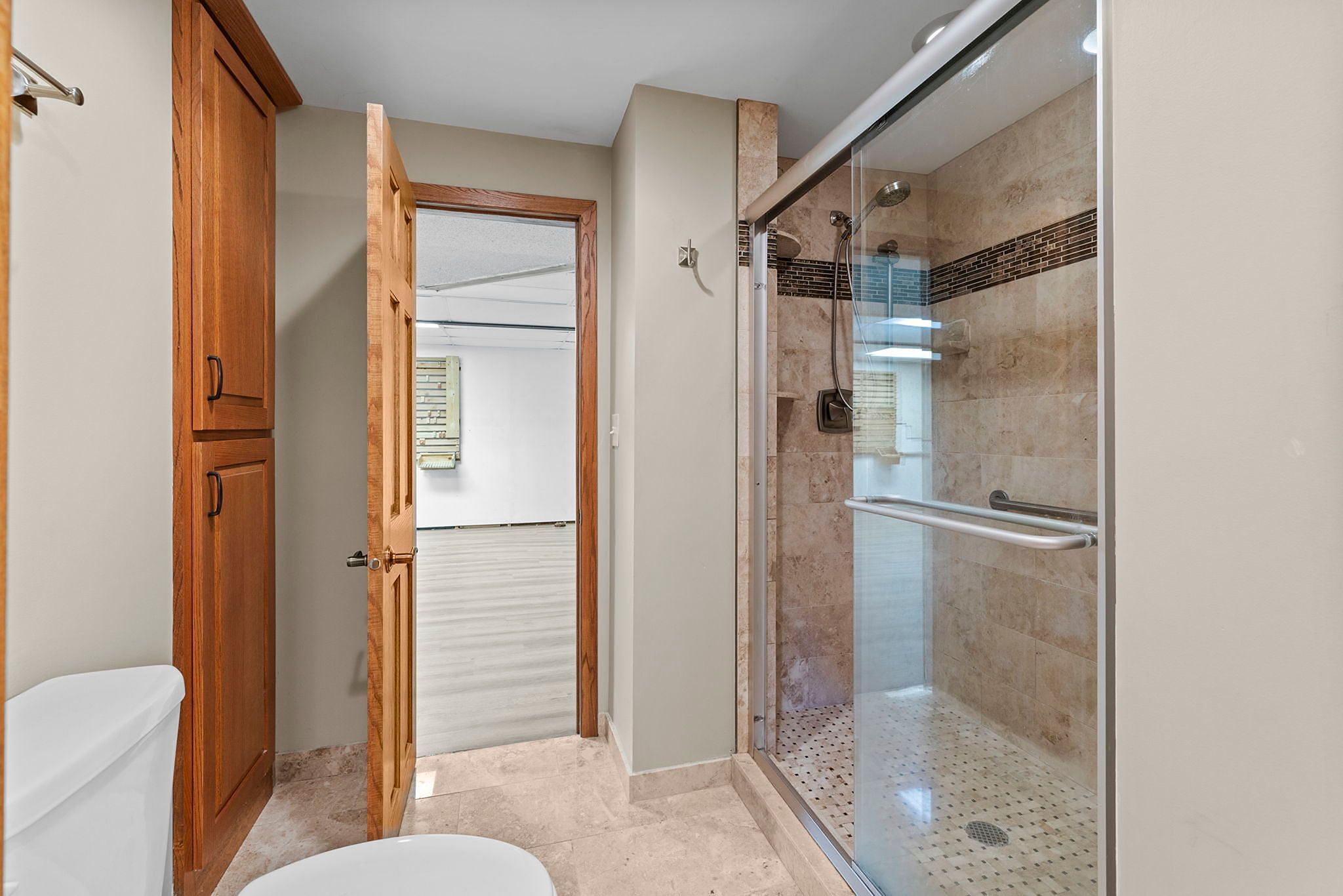 Modern bathroom with a glass-enclosed shower, tiled walls and floor, wooden cabinet, and toilet. An open wooden door leads to an adjacent room with light-colored flooring.