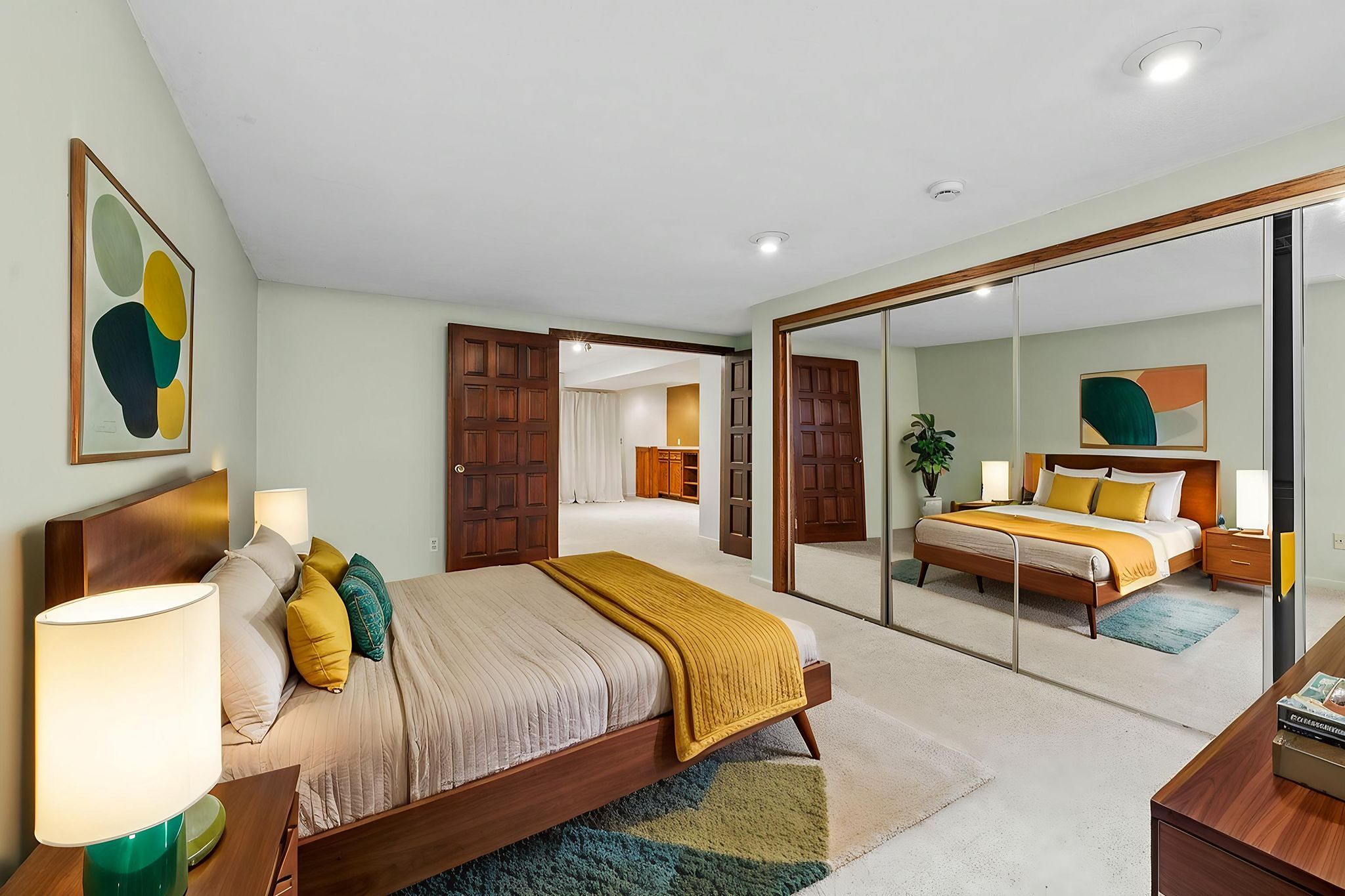Spacious bedroom with a wooden bed, yellow and green accents, mirrored closet doors, modern lamps, and abstract wall art. The room connects to another area through double wooden doors.