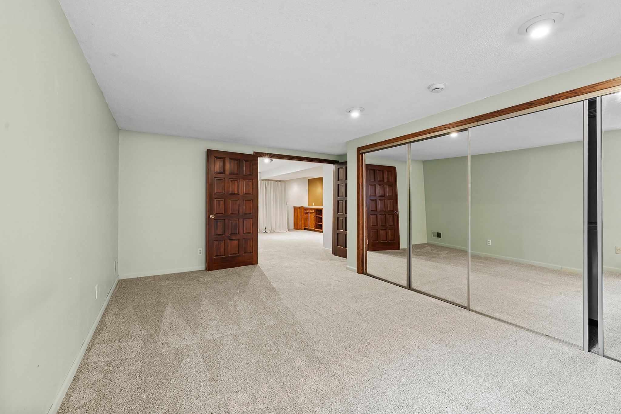 Spacious carpeted room with soft green walls, wooden double doors, and a large mirrored closet. Recessed ceiling lights brighten the area, and another room with cabinetry is visible through the open doors.