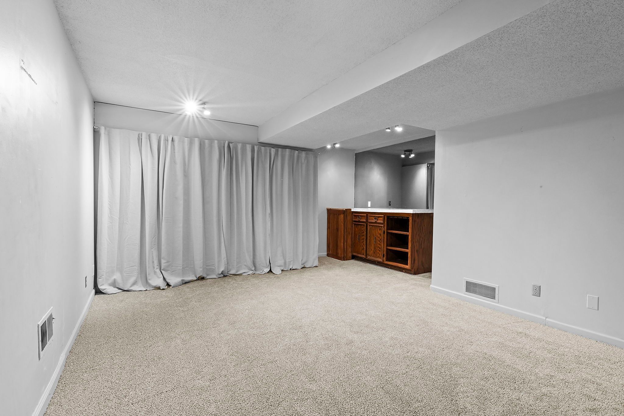 Basement room with beige carpet, white walls, closed gray curtains, and a wooden bar area with shelves and drawers under a lowered ceiling section. Bright overhead lights illuminate the space.