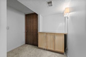 A small, carpeted room with white walls, a wooden door, a light wood cabinet, and a wall-mounted light fixture above the cabinet. An air vent is visible on the ceiling.