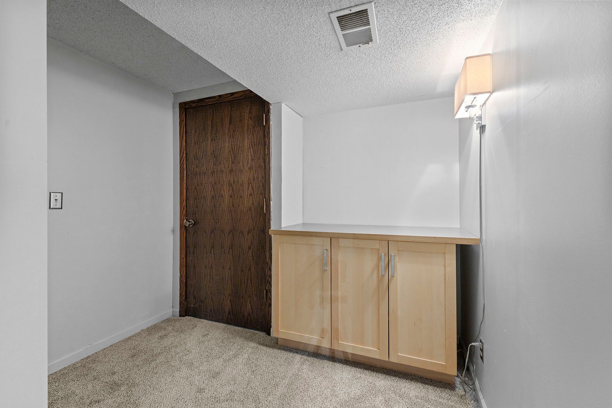 A small, carpeted room with white walls, a wooden door, a light wood cabinet, and a wall-mounted light fixture above the cabinet. An air vent is visible on the ceiling.