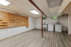 Unfinished basement room with light wood flooring, washer and dryer in the corner, laundry sink, fluorescent ceiling lights, and a wooden storage rack on one wall. Two small windows bring in some natural light.