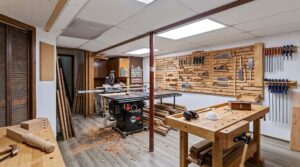 A well-organized woodworking shop with wooden workbenches, tools mounted on the walls, wood pieces stacked, a table saw, clamps, and safety gear on the bench. Wood shavings are scattered on the floor.