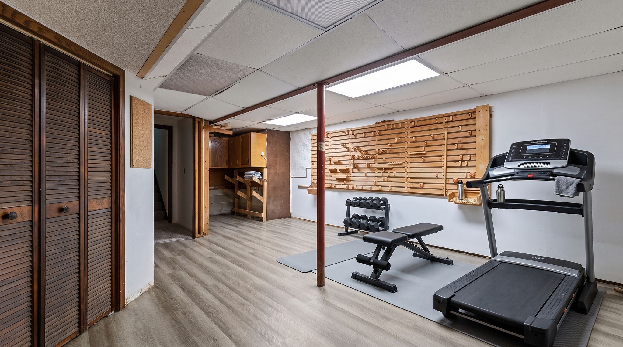 Home gym with treadmill, weight bench, dumbbells, and wooden slat wall. Room has light wood floors, white ceiling tiles, and a closet with louvered doors. Stairs are visible in the background.