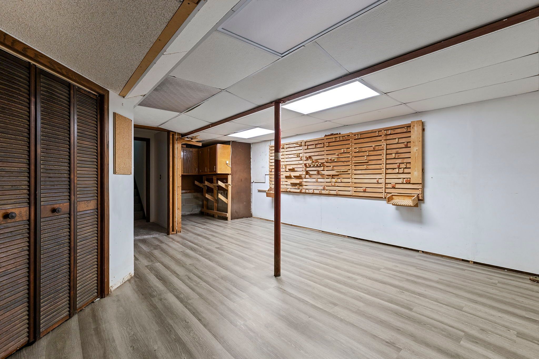 Unfurnished basement room with wood laminate flooring, fluorescent ceiling lights, wood closet doors, a support pole, and a large pegboard organizer mounted on a white wall. A storage or workshop area is visible in the back.