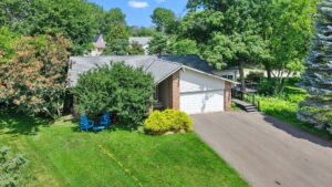 A suburban house with a two-car garage, surrounded by trees and greenery. A paved driveway leads to the garage, and two blue chairs sit on the front lawn near bushes.