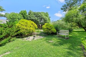 A white wooden bench sits on a grassy lawn near a small stone-bordered garden, surrounded by lush green trees and shrubs under a bright, sunny sky.
