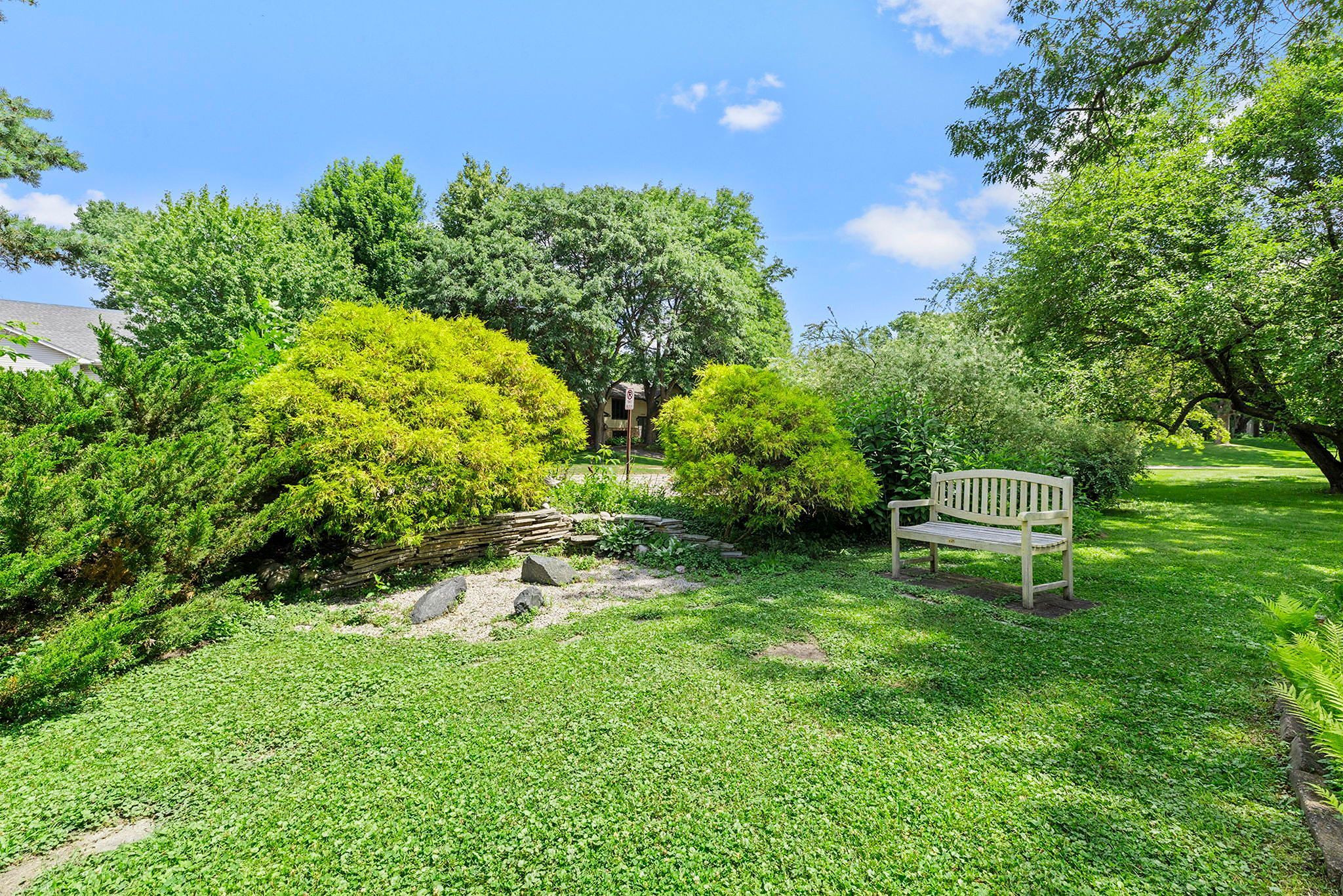 A white wooden bench sits on a grassy lawn near a small stone-bordered garden, surrounded by lush green trees and shrubs under a bright, sunny sky.