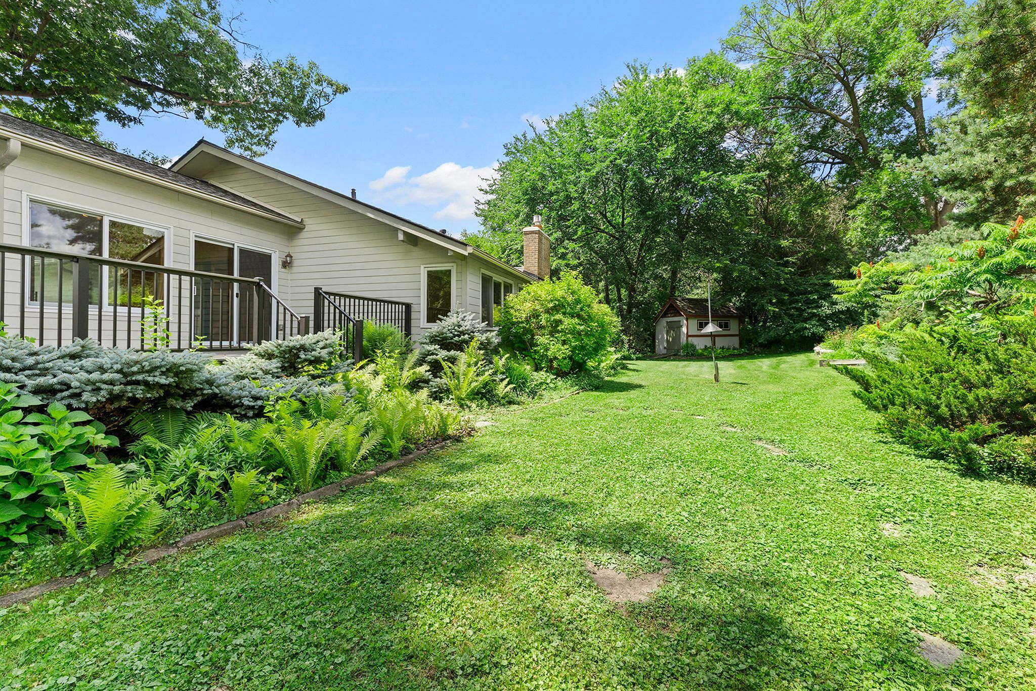 Backyard with green grass, lush landscaping, a white house with a deck on the left, and a small shed or gazebo in the background surrounded by tall trees and shrubs under a blue sky.