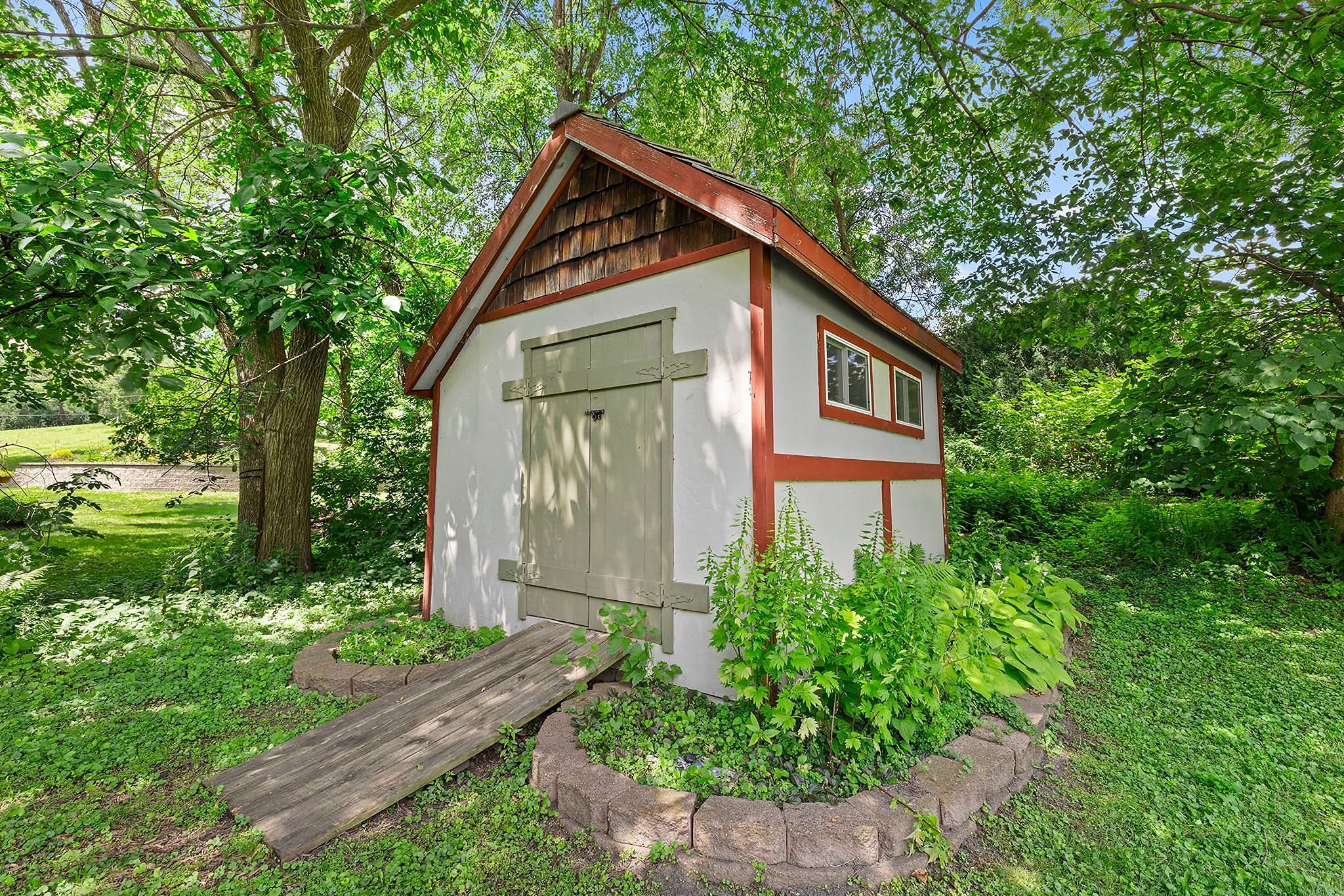 Small garden shed with beige doors, red trim, and a ramp, surrounded by green plants, under large trees with abundant foliage on a sunny day.
