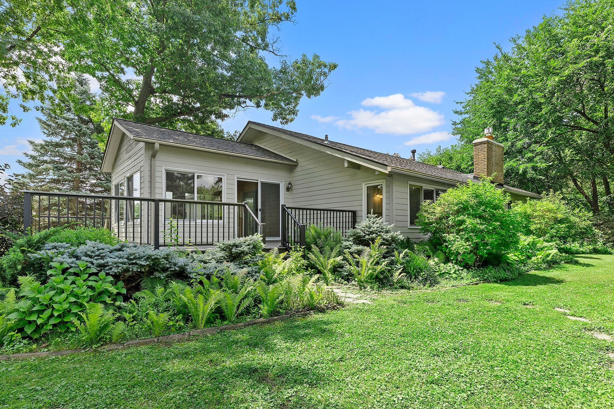 Single-story house with gray siding, large windows, and a back porch with railings. Lush green plants and bushes line the yard, with trees in the background under a bright blue sky with scattered clouds.