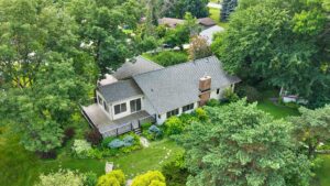 Aerial view of a house surrounded by lush green trees and landscaping, with a lawn, garden path, wooden deck, and nearby neighborhood homes visible.
