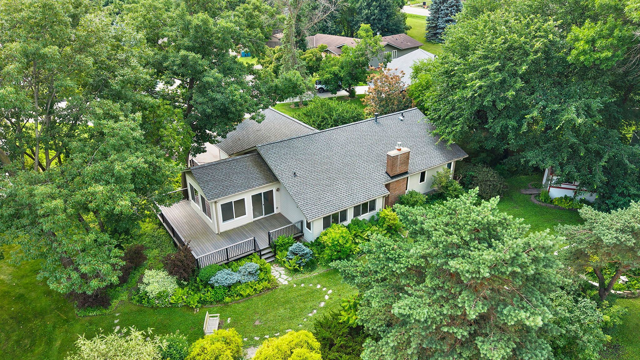 Aerial view of a house surrounded by lush green trees and landscaping, with a lawn, garden path, wooden deck, and nearby neighborhood homes visible.