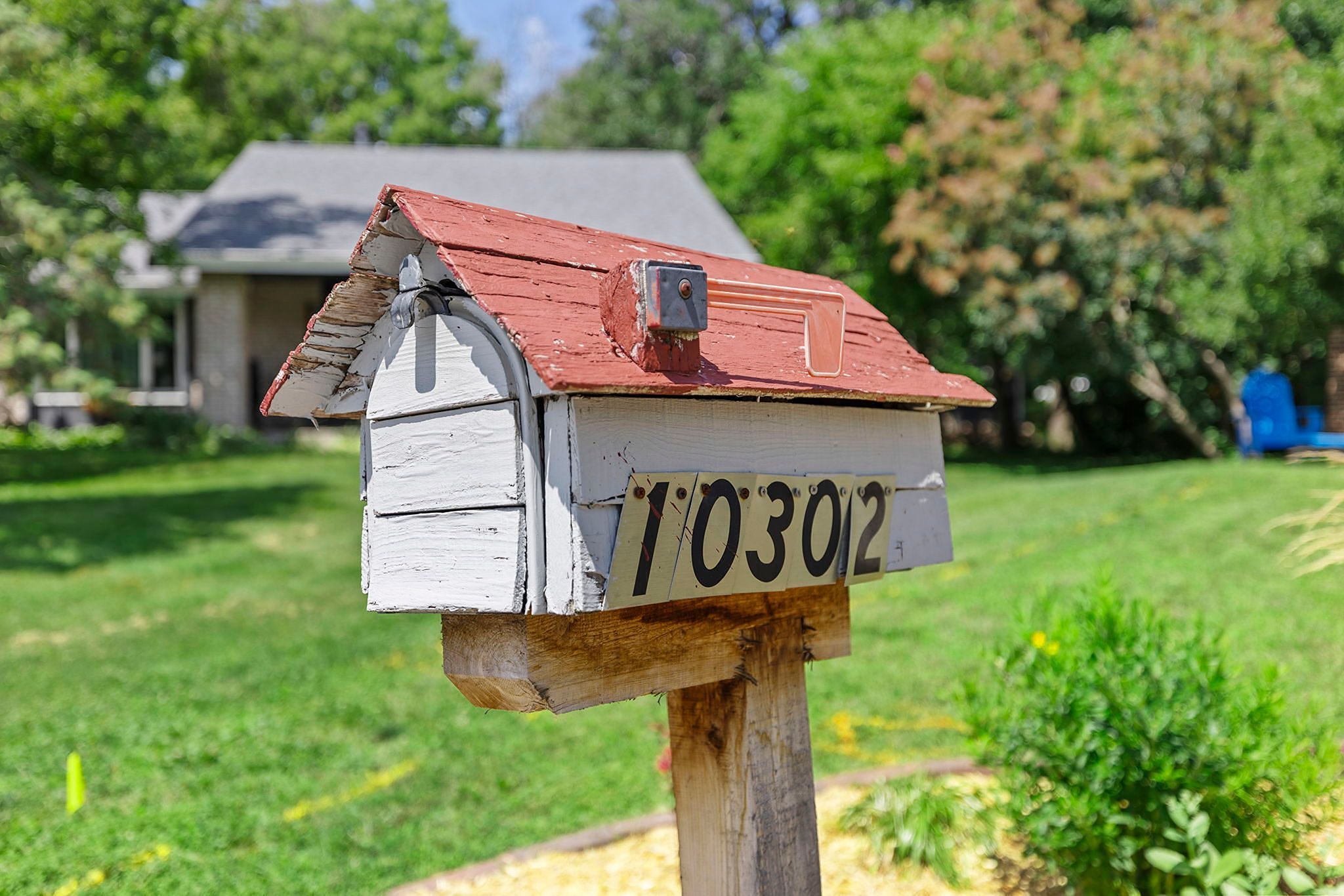 A white mailbox with a red roof and the number 10302 is mounted on a wooden post in a sunny yard, with a house and green trees in the background.