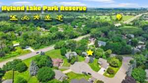 Aerial view of a suburban neighborhood near Hyland Lake Park Reserve, with icons for swimming, biking, hiking, running, and skiing. A yellow star and pin highlight specific houses and locations. Lush trees and green spaces surround the area.